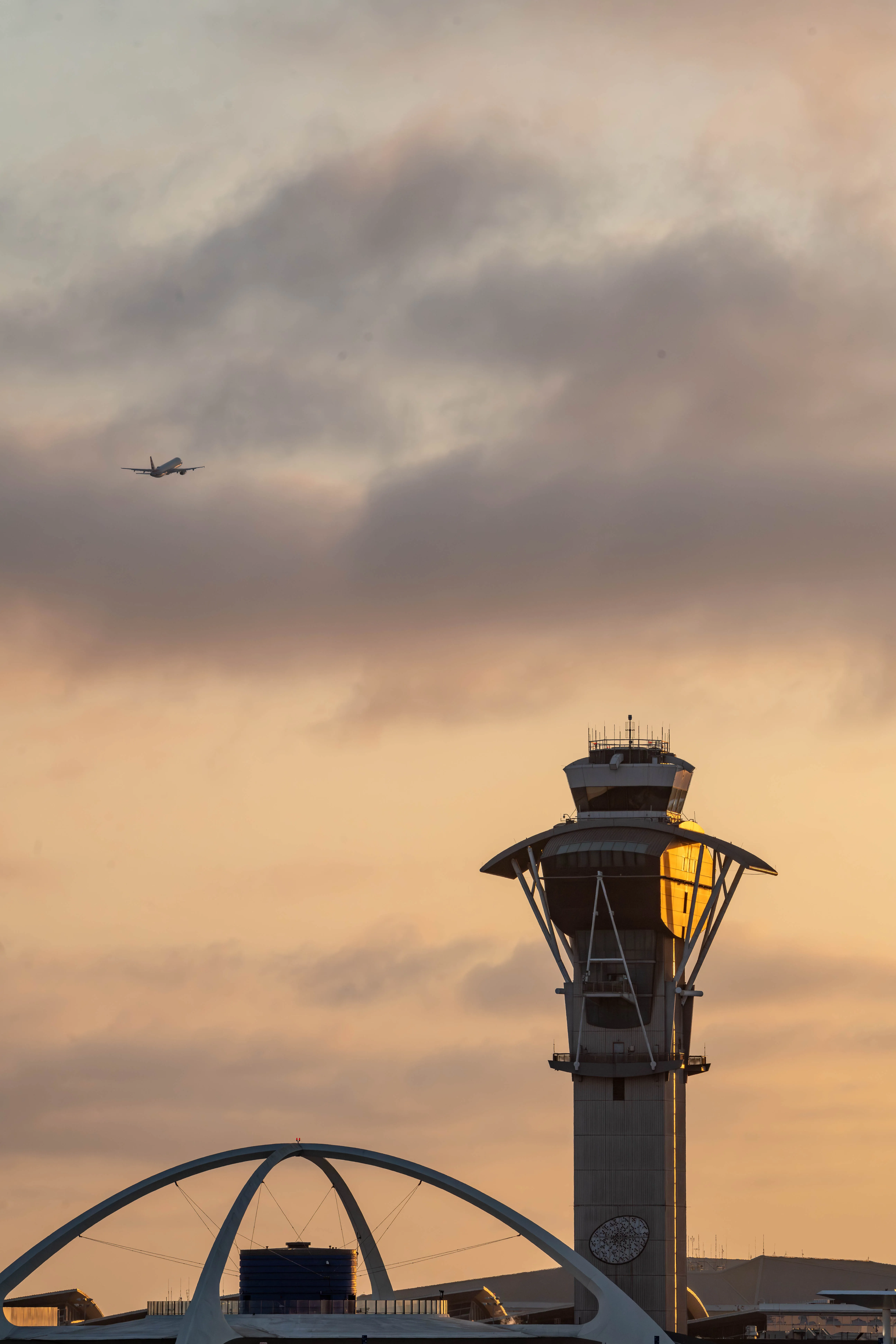Mid-century modern architecture photography by Thomas McClure of the LAX Theme Building & control tower in Los Angeles glowing at twilight. Airplane flies off in backgroud.